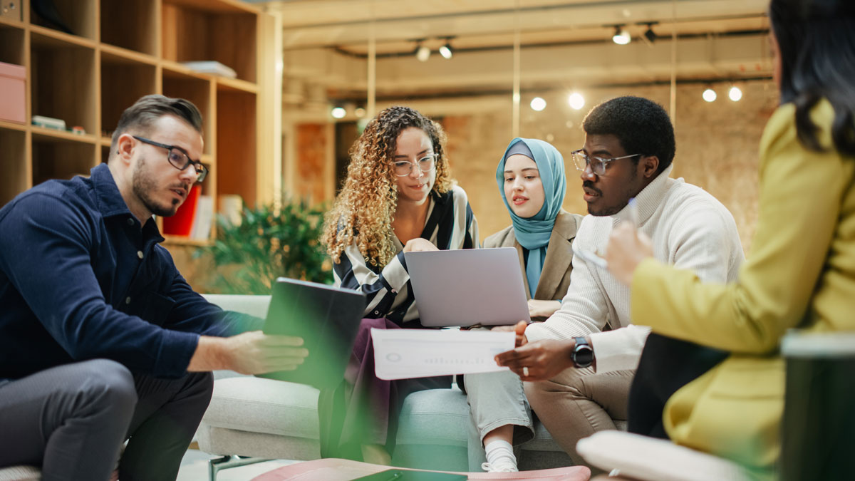 A Group of Multiethnic People Brainstorming Using Laptop and Charts in a Meeting Room at Office. Enthusiastic Young Adults Analyzing New Data To Improve Startup Performance. Medium Shot