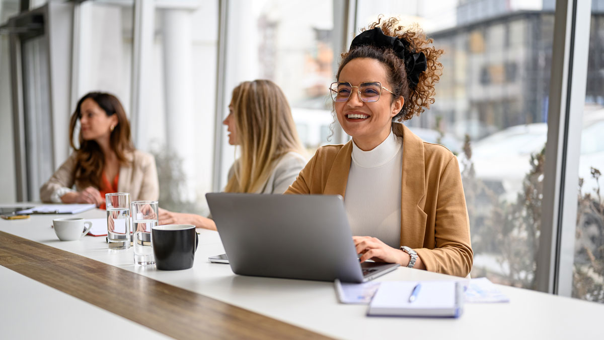 Happy businesswoman in eyeglasses working over laptop while siting with colleagues in modern board room