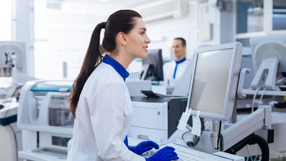 Nice touch. Energetic positive female laboratory assistant looking at the screen, typing while standing near the medical equipment