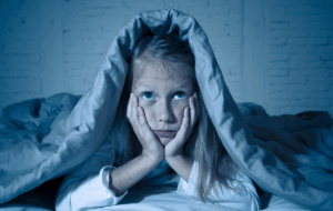 Das süße kleine, kaukasische Mädchen, das im Bett liegt und mit einer Decke bedeckt ist, die sich erschöpft und schlaflos an Schlaflosigkeit Depression Leid in Kinder Emotionale und Schlafstörungen Konzept.