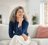 Glücklich entspannt 50 Jahre alte Frau sitzt auf Sofa zu Hause wegschauend. Lächelnde Frau mittleren Alters posiert im Wohnzimmer zu Hause. Reife ältere Frau mit lockigen grauen Haar entspannen auf Couch genießen Chill