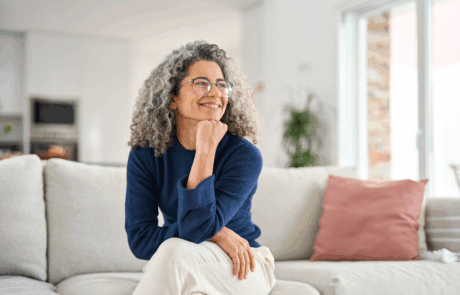 Glücklich entspannt 50 Jahre alte Frau sitzt auf Sofa zu Hause wegschauend. Lächelnde Frau mittleren Alters posiert im Wohnzimmer zu Hause. Reife ältere Frau mit lockigen grauen Haar entspannen auf Couch genießen Chill