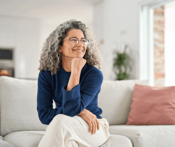 Glücklich entspannt 50 Jahre alte Frau sitzt auf Sofa zu Hause wegschauend. Lächelnde Frau mittleren Alters posiert im Wohnzimmer zu Hause. Reife ältere Frau mit lockigen grauen Haar entspannen auf Couch genießen Chill