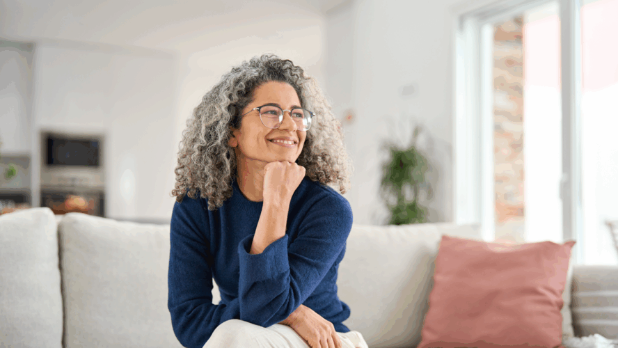 Glücklich entspannt 50 Jahre alte Frau sitzt auf Sofa zu Hause wegschauend. Lächelnde Frau mittleren Alters posiert im Wohnzimmer zu Hause. Reife ältere Frau mit lockigen grauen Haar entspannen auf Couch genießen Chill