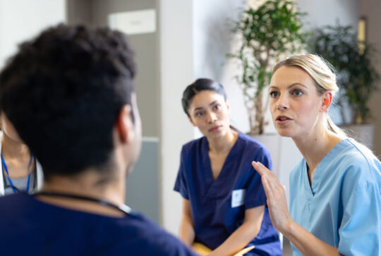 Caucasian female doctor sitting talking with diverse colleagues at hospital