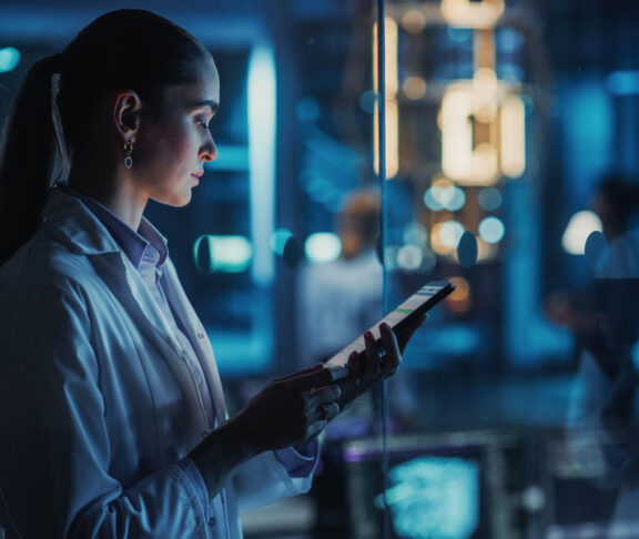 Female Scientist Using a Tablet to Monitor the Work of a Quantum Computer, Focusing on Data Flow, Compute and Hardware Stability. Futuristic Dark Setting with Advanced Technological Office