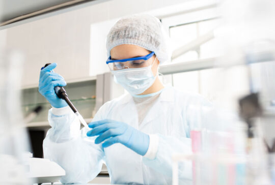 Serious concentrated female microbiologist in sterile clothing and safety goggles sitting at table and dropping reagent in petri dish while doing research in laboratory