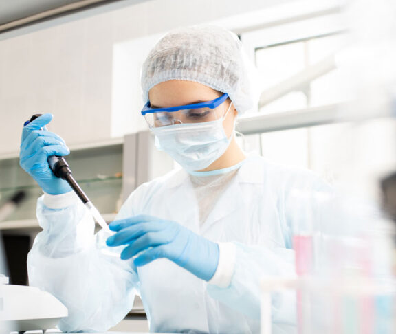 Serious concentrated female microbiologist in sterile clothing and safety goggles sitting at table and dropping reagent in petri dish while doing research in laboratory