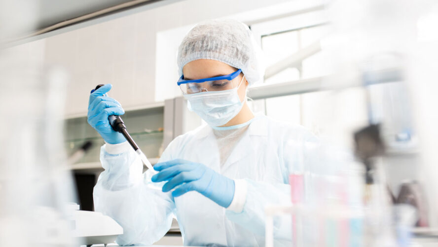Serious concentrated female microbiologist in sterile clothing and safety goggles sitting at table and dropping reagent in petri dish while doing research in laboratory