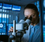 Female Medical Research Scientist Looking Through the Microscope Types Acquired Data in the Computer. Laboratory. In the Laboratory with Multiple Screens Showing MRI / CT Brain Scan Images