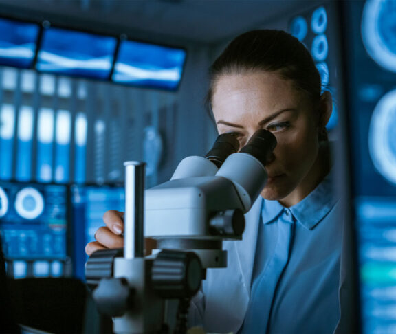 Female Medical Research Scientist Looking Through the Microscope Types Acquired Data in the Computer. Laboratory. In the Laboratory with Multiple Screens Showing MRI / CT Brain Scan Images