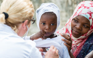 Female Caucasian doctor listening heart beat and breathing of little African girl with stethoscope.Mother holding the child