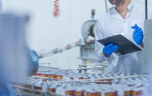 A food processing supervisor wearing protective gear and holding a clipboard, inspecting machinery on a production line, emphasizing quality control, safety, and efficiency in industrial operations
