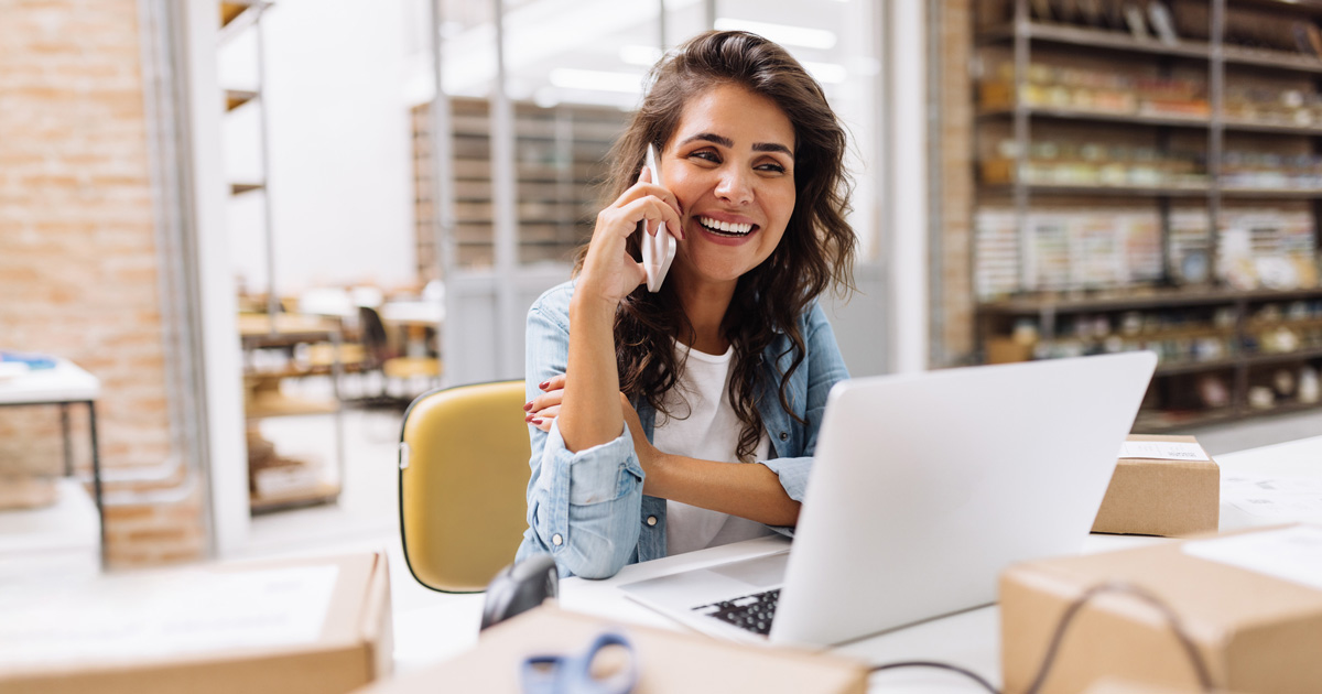 Happy young businesswoman speaking on the phone while working in a warehouse. Online store owner making plans for product shipping. Creative female entrepreneur running an e-commerce small business.