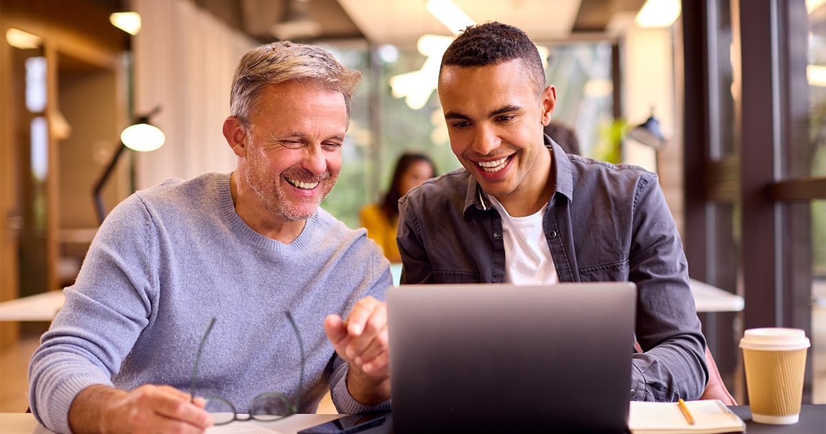 Mature Businessman Mentoring Younger Colleague Working On Laptop At Desk