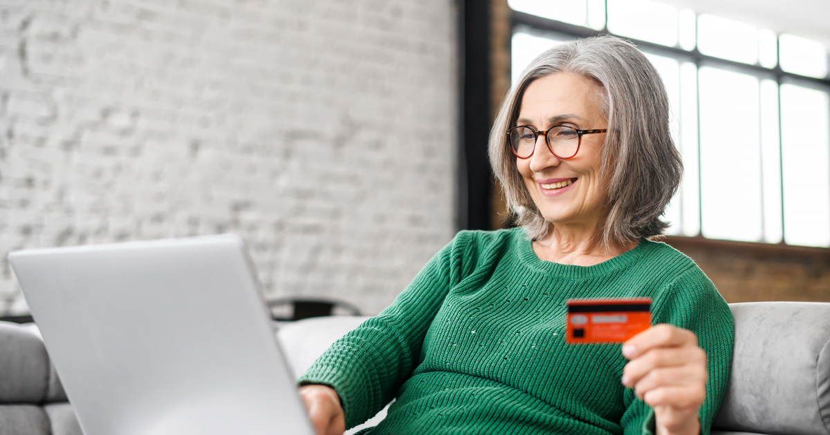Pretty senior businesswoman in glasses and green jumper sitting at the desk using laptop and credit card for online payment from home, typing number to enter internet banking account, ordering food