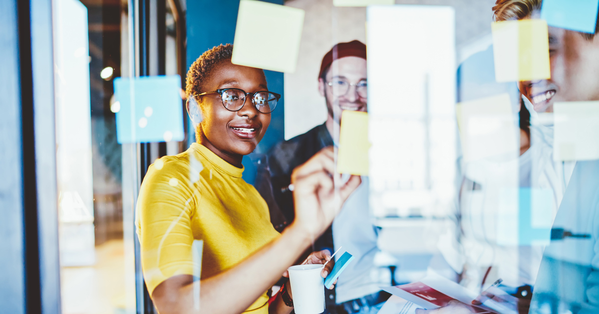 Group of happy hipster guys making notes on colorful memo sticks at glass wall while creating interesting ideas, cheerful multiracial male and female smiling while working with startup project