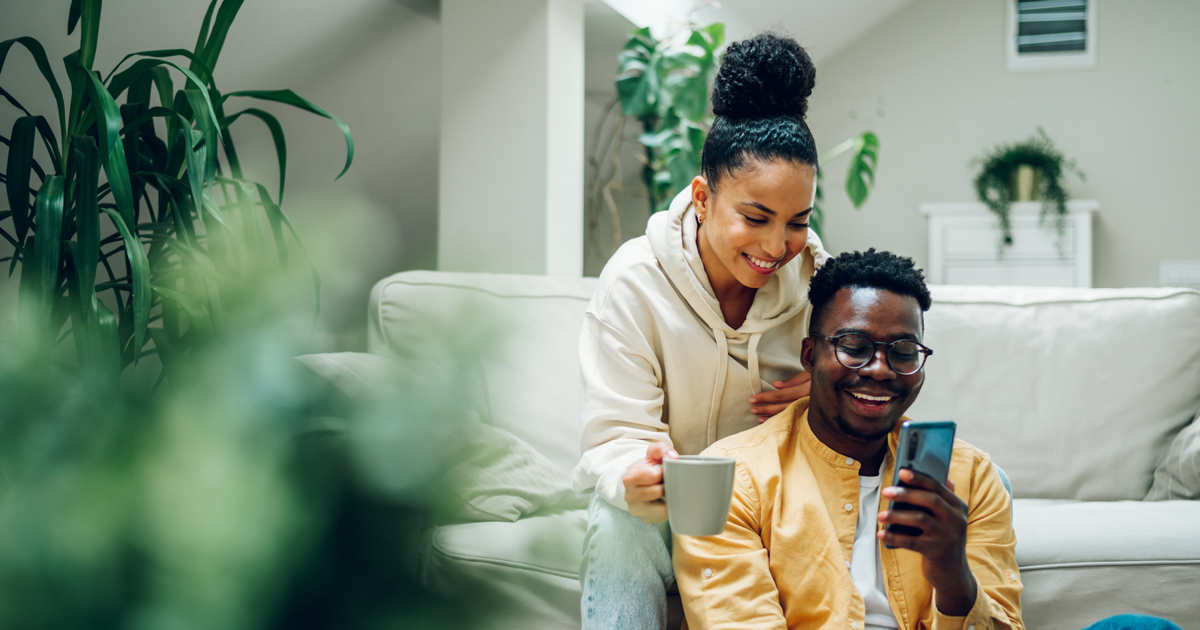 Multiracial young couple sitting on a comfortable sofa using smartphone and holding cups of hot beverages. Spouse relaxing together on cozy couch and online shopping on a mobile phone. Copy space.