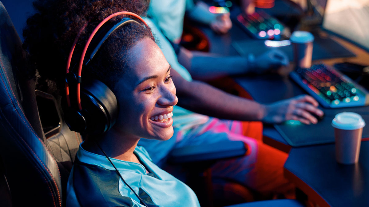 Cheerful African female cybersport gamer wearing wired headset sitting on gaming chair and watching game of teammate in cyber club