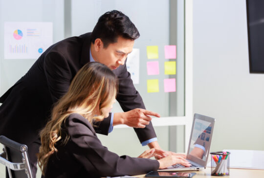 Millennial Asian successful professional male businessman mentor in formal suit coaching training helping female businesswoman secretary trainee working with laptop computer in office meeting room.