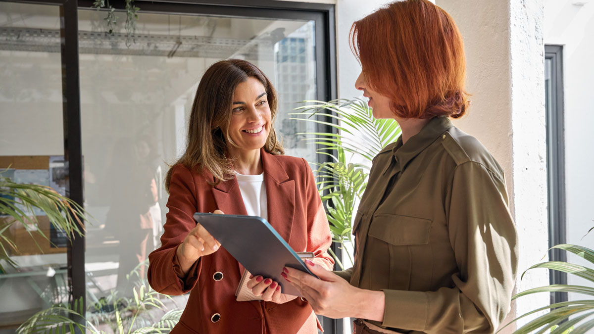 Two happy business women of young and middle age talking standing in creative green office space. Smiling professional ladies employees colleagues having conversation using digital tablet at work