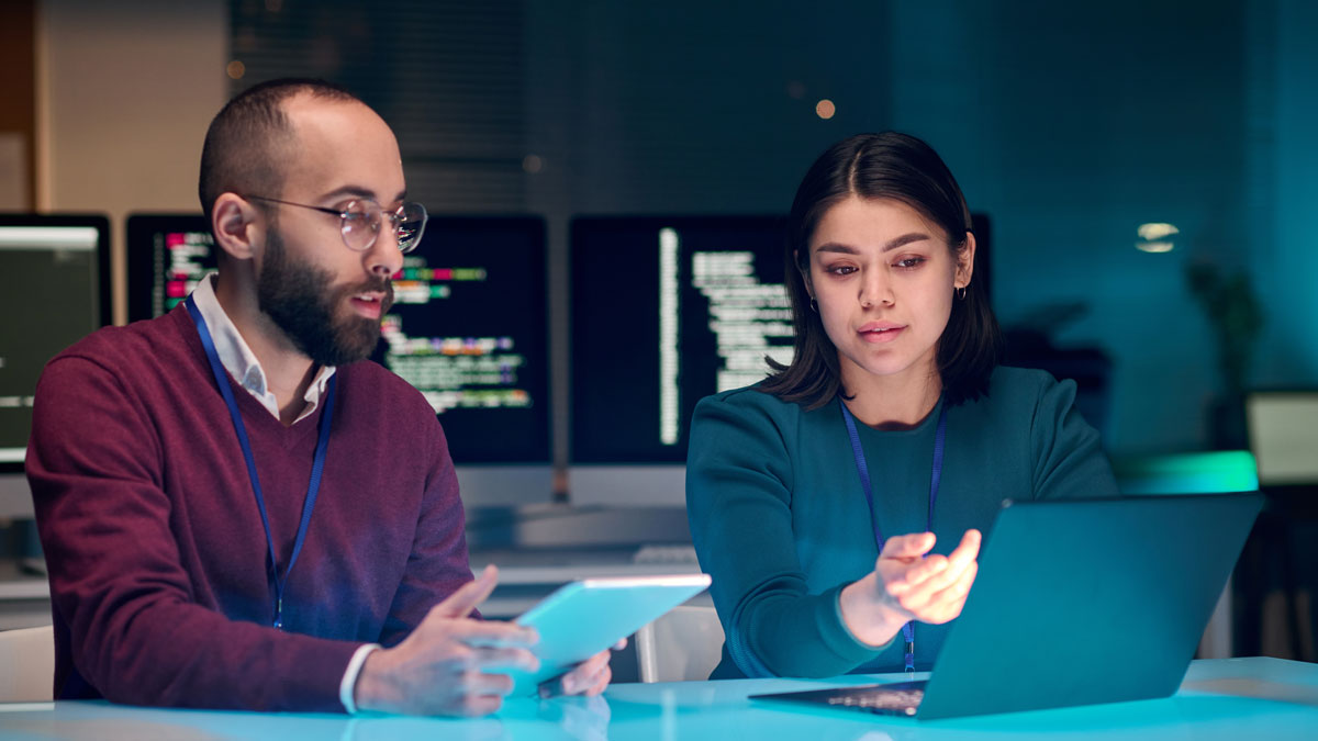 Portrait of young woman as female IT security expert using laptop with male colleague in blue neon light