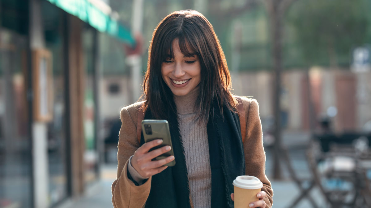 Pretty young woman using her mobile phone while drinking cup of coffee walking the streets of the city.