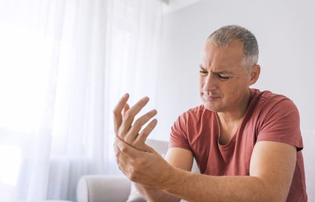 Photo of Mature man suffering from wrist pain at home while sitting on sofa during the day. Clenched painful hands
