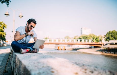 Staying ahead with new technologies.Hipster sits by the river in looking at tablet.He drinks coffee.Next to him is his longboard.He enjoys a nice summer day