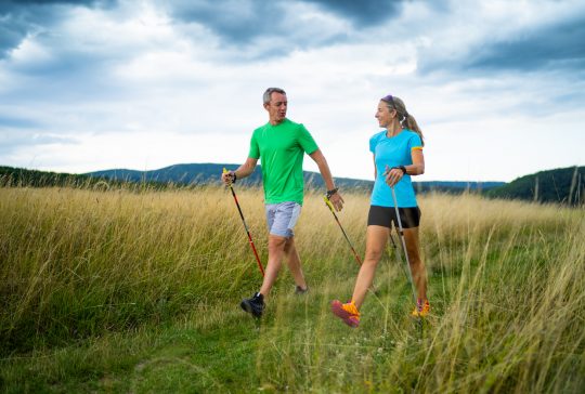 active smiling middle aged couple doing nordic walking sport in grassland with shallow focus cloudy overcast sky dark clouds front view
