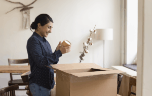 Fröhliches junges indisches Jungpferd packt Sachen zum Verlassen der Wohnung. Kunde, der ein Paket mit Einkauf im Webshop, Öffnen des Kartons, Papiercontainer, Lächeln, Spaß am Laufen erhält