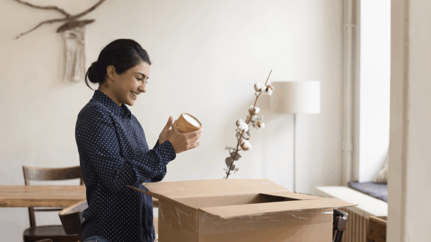 Fröhliches junges indisches Jungpferd packt Sachen zum Verlassen der Wohnung. Kunde, der ein Paket mit Einkauf im Webshop, Öffnen des Kartons, Papiercontainer, Lächeln, Spaß am Laufen erhält