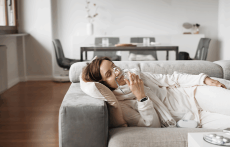Gelassene Frau atmet durch Sauerstoffmaske und mit Sauerstoffkonzentrator für die Atemtherapie zu Hause, finden Komfort und Erleichterung auf ihrem Wohnzimmersofa