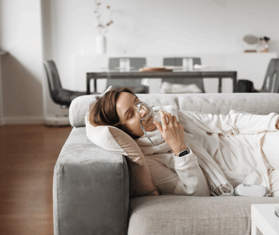 Gelassene Frau atmet durch Sauerstoffmaske und mit Sauerstoffkonzentrator für die Atemtherapie zu Hause, finden Komfort und Erleichterung auf ihrem Wohnzimmersofa