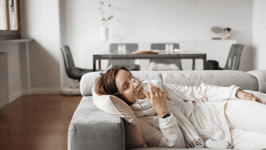 Gelassene Frau atmet durch Sauerstoffmaske und mit Sauerstoffkonzentrator für die Atemtherapie zu Hause, finden Komfort und Erleichterung auf ihrem Wohnzimmersofa