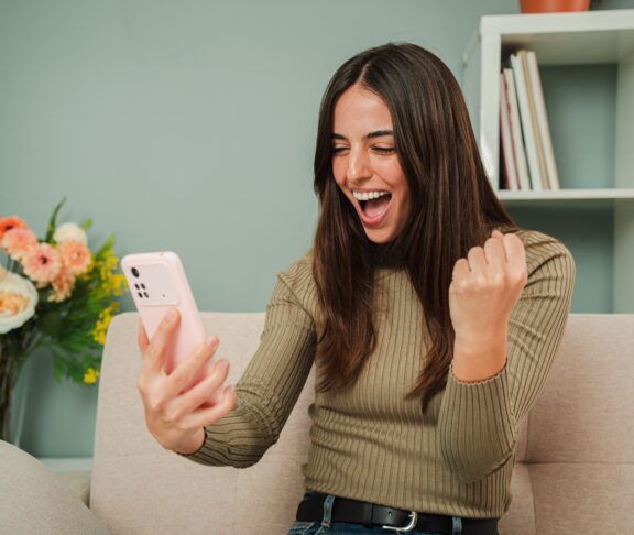 Frau feiert, dass sie eine Sportwette gewonnen hat, während sie das Ergebnis auf einem Smartphone-App. Glückliche Wetten für Frauen in einem Online-Casino mit einem Handy. Eine aufgeregte Dame, die den Lottogewinn sieht