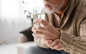 Schnittbild eines alten Mannes mit Parkinson-System, das ein Glas Wasser in zitternden Händen hält, während er zu Hause sitzt
