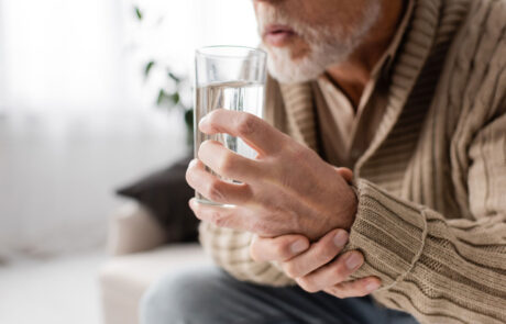 Schnittbild eines alten Mannes mit Parkinson-System, das ein Glas Wasser in zitternden Händen hält, während er zu Hause sitzt