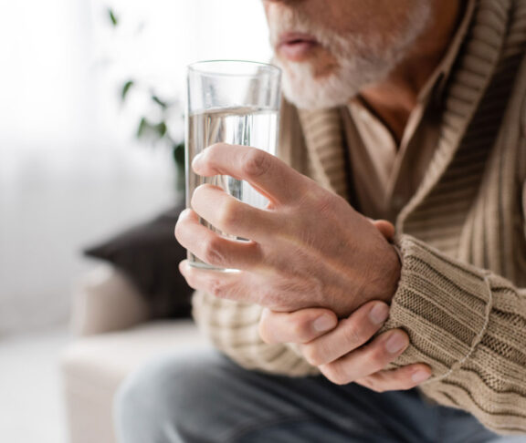 Schnittbild eines alten Mannes mit Parkinson-System, das ein Glas Wasser in zitternden Händen hält, während er zu Hause sitzt