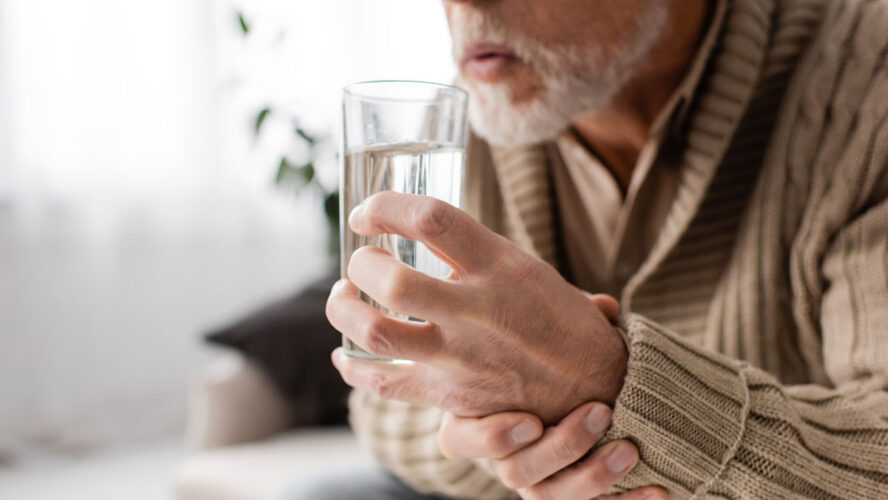 Schnittbild eines alten Mannes mit Parkinson-System, das ein Glas Wasser in zitternden Händen hält, während er zu Hause sitzt