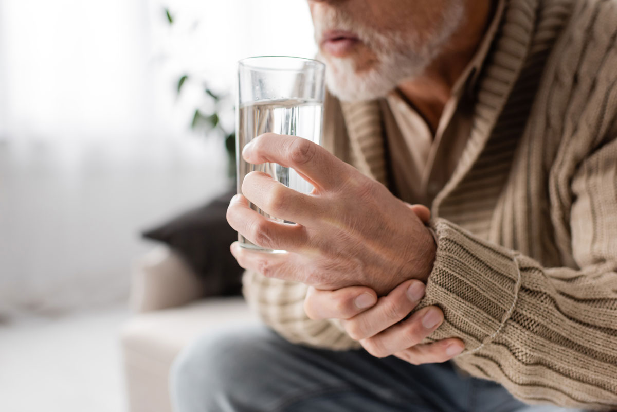 Schnittbild eines alten Mannes mit Parkinson-System, das ein Glas Wasser in zitternden Händen hält, während er zu Hause sitzt