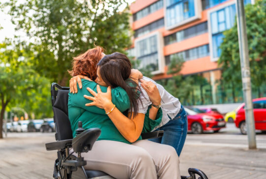 Zarter Moment einer kaukasischen Frau mit Behinderung, die ihre asiatische Betreuerin in der Stadt umarmt