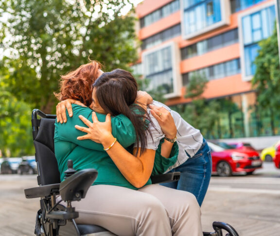 Zarter Moment einer kaukasischen Frau mit Behinderung, die ihre asiatische Betreuerin in der Stadt umarmt