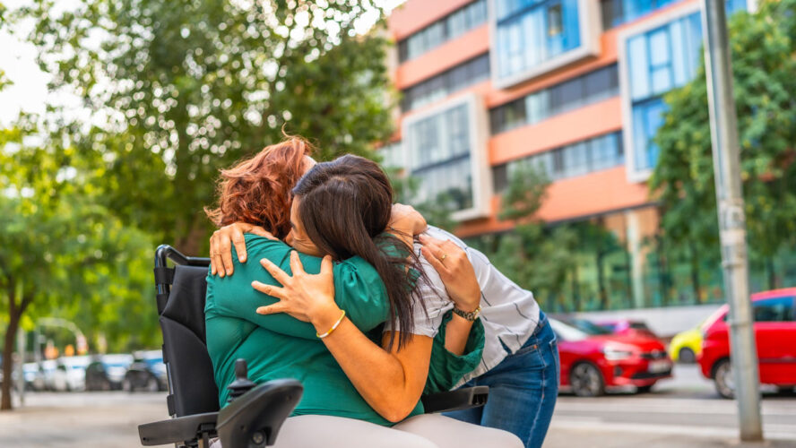 Zarter Moment einer kaukasischen Frau mit Behinderung, die ihre asiatische Betreuerin in der Stadt umarmt