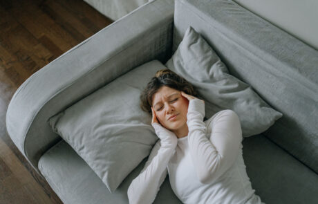 Junge Frau mit starken Kopfschmerzen, die zu Hause auf dem Sofa liegt, die Schläfen mit den Fingern berührt, die Augen geschlossen hält, sich ungesund fühlt, Migräne oder Stress erlebt