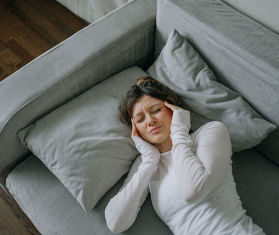 Junge Frau mit starken Kopfschmerzen, die zu Hause auf dem Sofa liegt, die Schläfen mit den Fingern berührt, die Augen geschlossen hält, sich ungesund fühlt, Migräne oder Stress erlebt