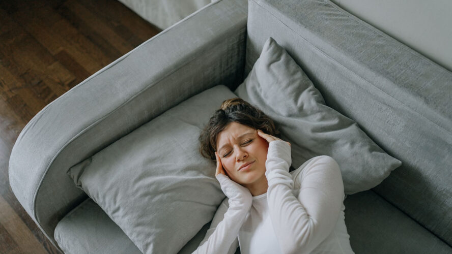 Junge Frau mit starken Kopfschmerzen, die zu Hause auf dem Sofa liegt, die Schläfen mit den Fingern berührt, die Augen geschlossen hält, sich ungesund fühlt, Migräne oder Stress erlebt