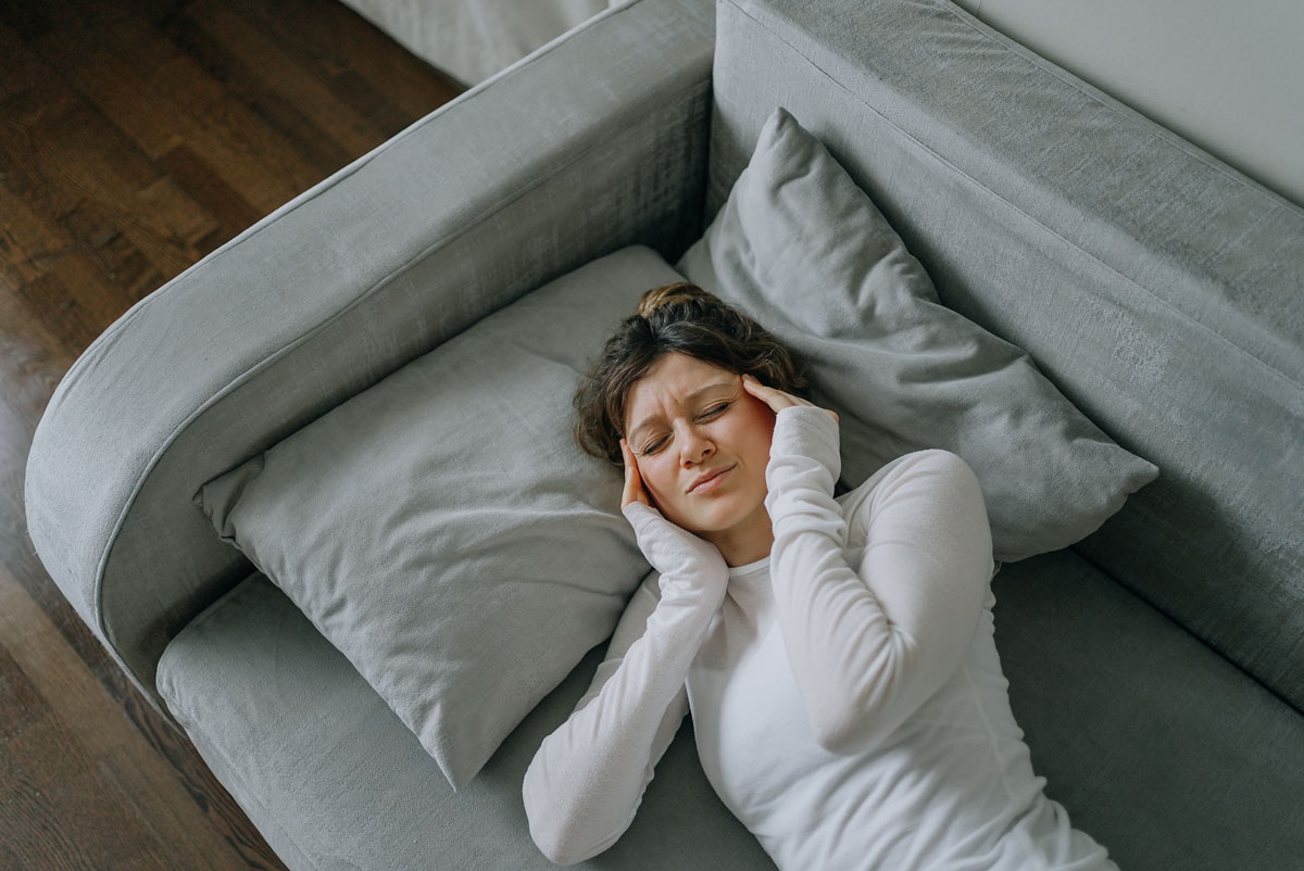 Junge Frau mit starken Kopfschmerzen, die zu Hause auf dem Sofa liegt, die Schläfen mit den Fingern berührt, die Augen geschlossen hält, sich ungesund fühlt, Migräne oder Stress erlebt