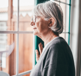 Alte ältere, grauhaarige Frau mit einer Brille, die am Fenster steht und nach draußen schaut. Senior Frau melancholische Träume zu Hause mit nachdenklichem Ausdruck.