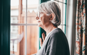 Alte ältere, grauhaarige Frau mit einer Brille, die am Fenster steht und nach draußen schaut. Senior Frau melancholische Träume zu Hause mit nachdenklichem Ausdruck.